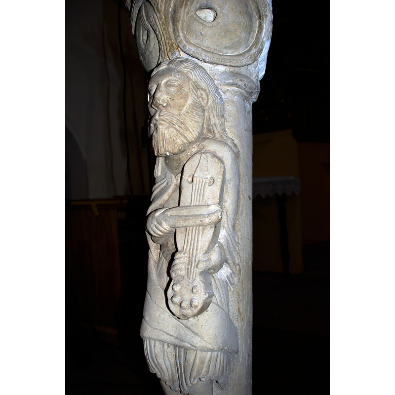 Source caption: "Estatua-columna en la iglesia de San Andrés de Revilla de Collazos (Palencia, Castilla y León). Actualemente hace función de atril y es la pieza escultórica más conocida del edificio. Alta de 1,20 m, se compone de basa sobre plinto de prominente toro inferior con una cabecita de felino a modo de garra, escocia lisa y toro superior atrofiado. Adosada al fuste aparece la figura de un tañedor de viola con arco, vestido con túnica cuyos fruncidos y ondulados pliegues aparecen bajo su sayón. Su rostro hierático luce larga cabellera, barba mediana y ojos almendrados e inexpresivos. Bien que sean evidentes las desproporciones del canon, éstas no restan gracia a la figura, que se despega airosa del fuste que la sustenta. El gran capitel que remata la columna se decora con dos series de tallos perlados vueltos y rematados por hojas que acogen cuatro gruesas bolas que pudieran querer representar frutos. Se ha supuesto esta columna perteneciente a la desaparecida ventana central del ábside. Sus dimensiones y el bisel de la parte posterior así lo sugieren, aunque la tipología de la basa y astrágalo no concuerda con los de las columnas conservadas en la ventana sur del hemiciclo. Se viene asimilando a este músico con el rey David, aunque falten elementos definitorios para determinar con certeza tal identificación."