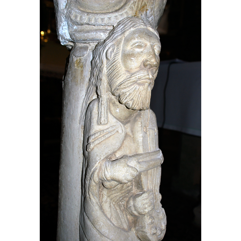 Source caption: "Estatua-columna en la iglesia de San Andrés de Revilla de Collazos (Palencia, Castilla y León). Actualemente hace función de atril y es la pieza escultórica más conocida del edificio. Alta de 1,20 m, se compone de basa sobre plinto de prominente toro inferior con una cabecita de felino a modo de garra, escocia lisa y toro superior atrofiado. Adosada al fuste aparece la figura de un tañedor de viola con arco, vestido con túnica cuyos fruncidos y ondulados pliegues aparecen bajo su sayón. Su rostro hierático luce larga cabellera, barba mediana y ojos almendrados e inexpresivos. Bien que sean evidentes las desproporciones del canon, éstas no restan gracia a la figura, que se despega airosa del fuste que la sustenta. El gran capitel que remata la columna se decora con dos series de tallos perlados vueltos y rematados por hojas que acogen cuatro gruesas bolas que pudieran querer representar frutos. Se ha supuesto esta columna perteneciente a la desaparecida ventana central del ábside. Sus dimensiones y el bisel de la parte posterior así lo sugieren, aunque la tipología de la basa y astrágalo no concuerda con los de las columnas conservadas en la ventana sur del hemiciclo. Se viene asimilando a este músico con el rey David, aunque falten elementos definitorios para determinar con certeza tal identificación."