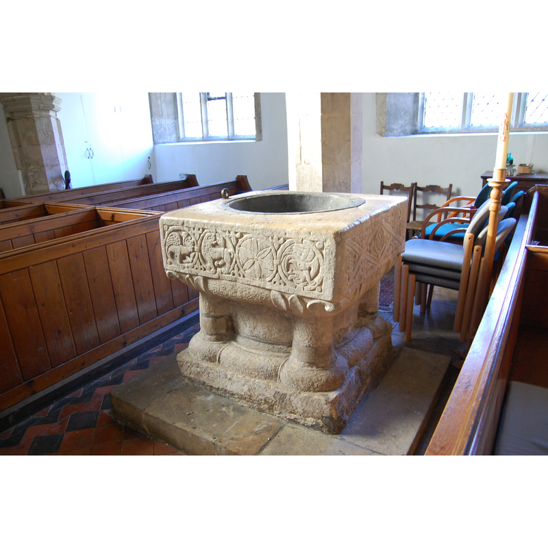 Source caption: "The church contains a remarkable 12th Century font with striking carvings on 3 sides including a head licking a leaf and three grotesque beasts."