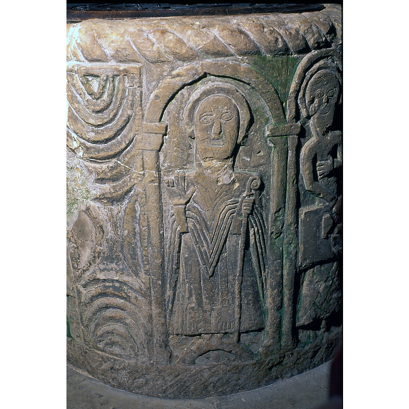 Priest in robe with stoles, giving sign of benediction. The priest is shown to the right of the Descent from the Cross. An ornamented panel is between the priest and the crucifixion scene.  Home (1908) suggests it may be St. Nicholas [cf. FontNotes]