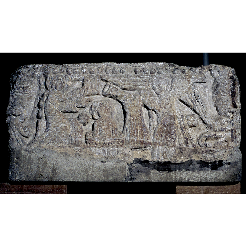 south side of the basin: John the Baptist (?), with a book in the left hand, while the right points to Christ; a star [of Bethlehem] appears between the left figure and Christ; in the centre of the scene is a square font (?) with an upper side decorated with a row of large beads, just like this font; in this font (?) is the upper body of a haloed figure; the carving is too crude and eroded to determine whether it is a child Christ or a grown-up; on the right side of the scene an angel with extended wings holds up the robes for Christ. Above the scene the upper end of the basin side is decorated with a row of large beads