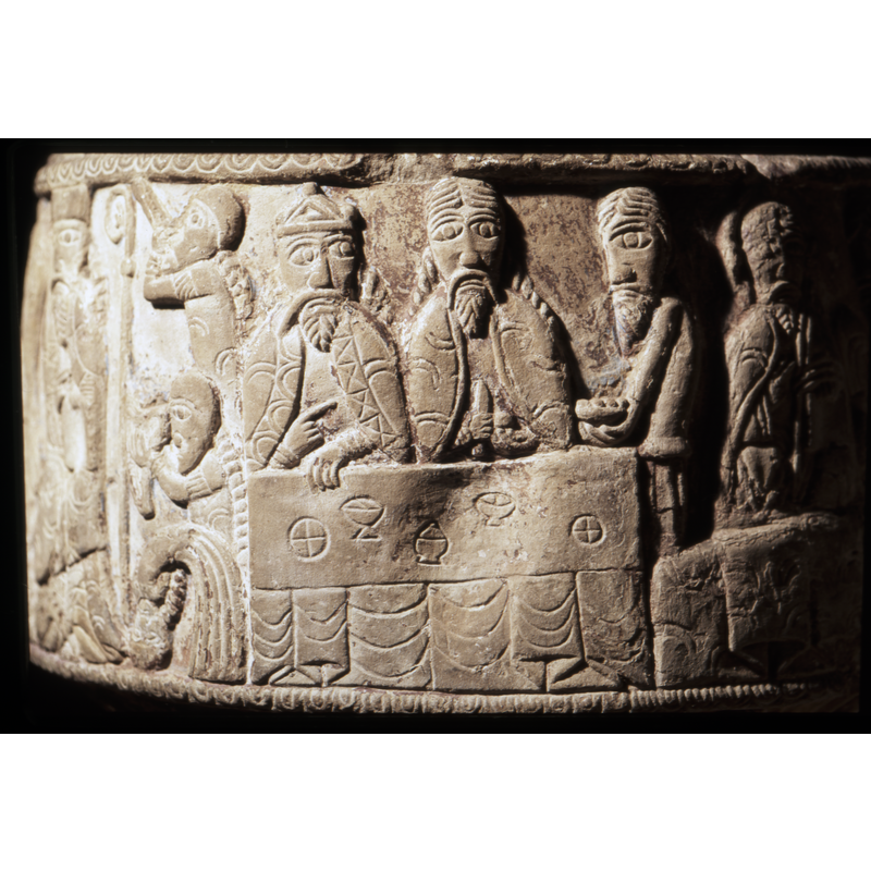 A table with bread and cups of wine. Two figures behind the table, both with long hair and braids and beards. The left figure has crown and looks to the figure on the right and points to it. The figure on the right, holds a knife in the right hand.To the right of the table is a figure in profile, bringing an object to the table