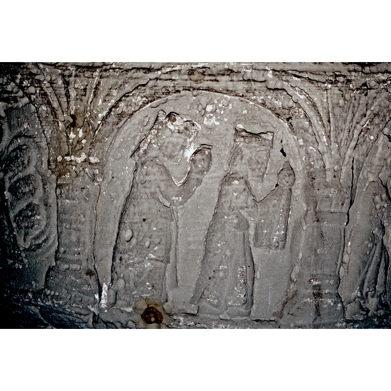 The scene takes place over two arches - the Adoration of the Magi.  In the first arch, to the right, the Madonna and Child, enthroned on Gotland throne, Mary haloed, full frontal, the right hand is visible and holding the child by the legs, The baby is in profile, haloed, his right hand points toward the magi. The figure to the left is the first magi, crowned, in profile, long robes, and is holding up something on a draped cloth over his hands. IN THE SECOND ARCH TO THE LEFT contains two magi, crowned, dressed similarily, these two hold their mantels with their left handsl. The figure to the right holds the gift on a cloth draped over his hands. - the other figure has no cloth draped over his hands.