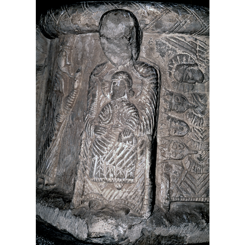 Madonna and Child. Madonna full frontal, face broken off; there is no halo, she has braids, the child sits on her lap, full frontal (check to see if there really are braids: yes, one to each side). Halo with cross on child, the right hand is raised to mother's shoulder in benediction, the left hand rests on a book on his left knee, she wears shoes, he does not. She holds him by the waist.
