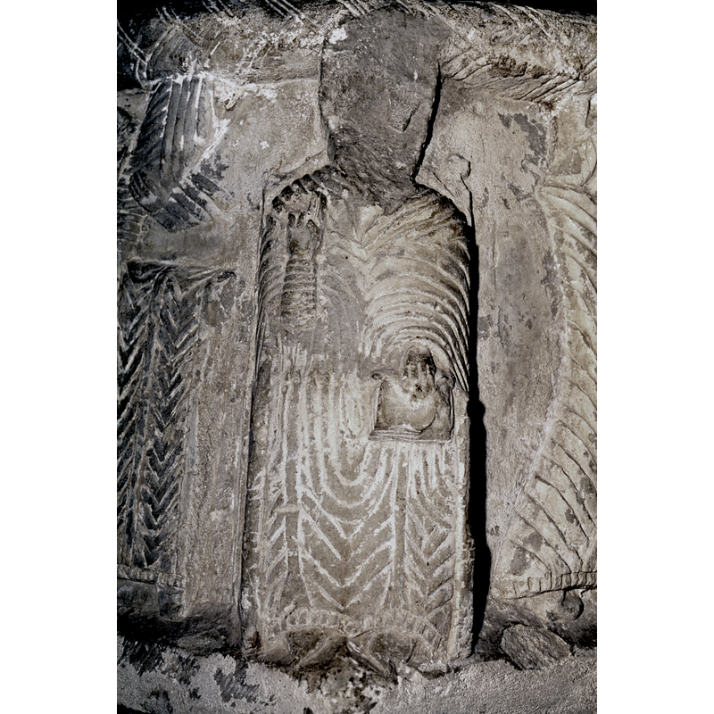 Bishop seated, enthroned, head is broken off; chair has an ornate back,unlike the Gotland chairs; right hand raised in benediction gesture, left hand holds a book in left knee.