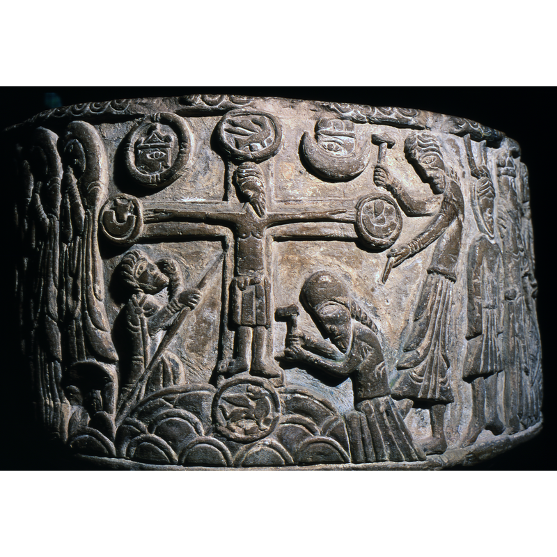 Crucifixion Scene. Christ on the cross, full front but head to the side, feet apart, wearing a long loin cloth to his knees. The cross has four medallions, one at each end, with the four Evangelist symbols. To the right two figures, one standing, the other kneeling, are driving nails into the cross. To the left, a kneeling figure holds a lance to Christ's body. To the left and right at the top of the cross, two symbols, Sun and Moon, with heads in them
