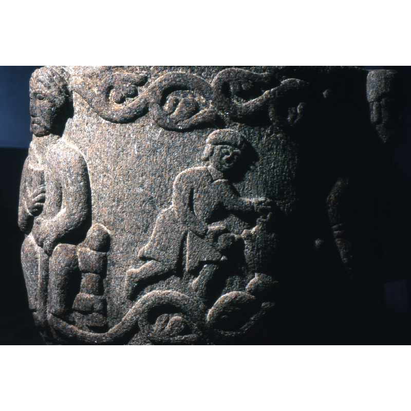 There is a figure in profile, semi-kneeling or walking, male, and wearing a flat hat, beret and a short tunic. The right hand holds a chalice like object, the left hand leans on a jug from which he is pouring liquid.