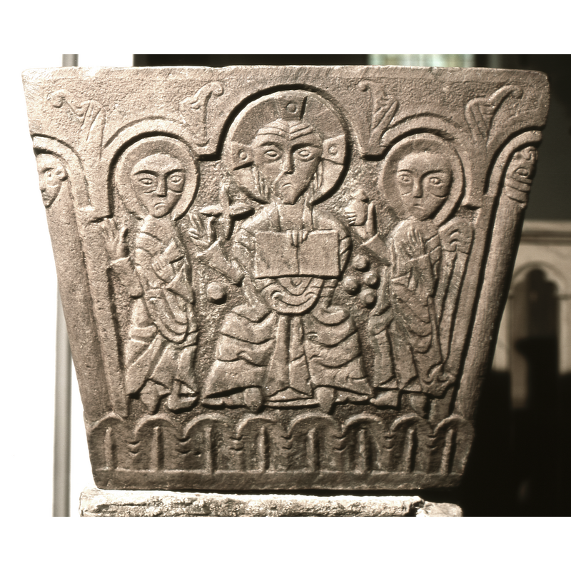 Christ enthroned. This is the central scene on the font and is framed by the three-arched arch on the fourth side of the font. Christ seated, holds an open book with his left hand and raises his right hand in benediction sign. To the left of his face is a 4-pointed star. To the right of him stands an angel holding a round container with a lid. (oil for baptism?) To the left of Christ is a second figure, haloed, holding a scroll (priest?). The corner columns on the font all have faces for capitals.