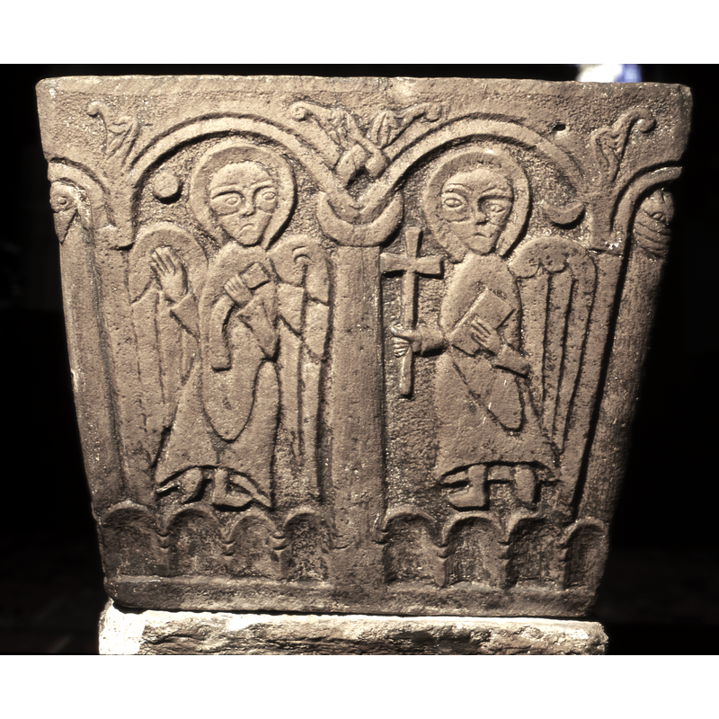 Two arches, the one on the left, has an angel both wings showing, right hand raised and left hand holds a scroll. The figure on the right, another angel, one wing showing, holds a cross in his right hand, the left hand holds a book. A moon is carved above his wing.