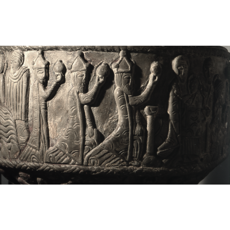 Adoration of the Magi. Moving from Left to Right. Three figures walking in profile, crowned, holding gifts. The first two are kneeling, resting their arm on knee. The last is unbearded, standing. The madonna and child to the right, enthroned, bench like throne, Mary not haloed, wraps the mantel around the child. Her right hand holds the child on her left side, hanging from her left wrist is a stole. Child in profile, right hand raised in benediction. Jesus is haloed with cross in haloe. To the right and behind, in the background a figure, probably female because of the headwear, stands in profile, hands in L-shape