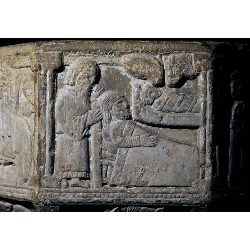 Nativity. Centre of one part of the program. No pictorial direction on this font - correct the earlier statement of Right to Left. To the left, Joseph, standing in profile, his left hand holds the left side of his face, the right hand holds the head post of the bed. He faces the right. Mary lies in bed, facing the right, the bed covers to her shoulders, the right hand rests on her abdomen. Above the bed and to the right of Mary's face, a manger with the child swaddled and facing the right. At the top right corner, the ox and mule.