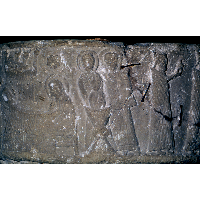 Christ's First Bath. The child is full frontal, emersed in font (chalice shape) - no hands visible. Being held from behind by the waist, by a woman (child has crossed haloe), Mary, haloed and head covered. To the right is another figure, haloed and no hands visible. (two women?) -- the cracked stone and the old rusty staples used in the repair are clearly visible here