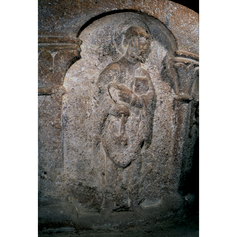 haloed; not clear whether the object in his right hand is a key or a censer; the book is held in the crook of the left arm while the hand points to the left his extended index finger